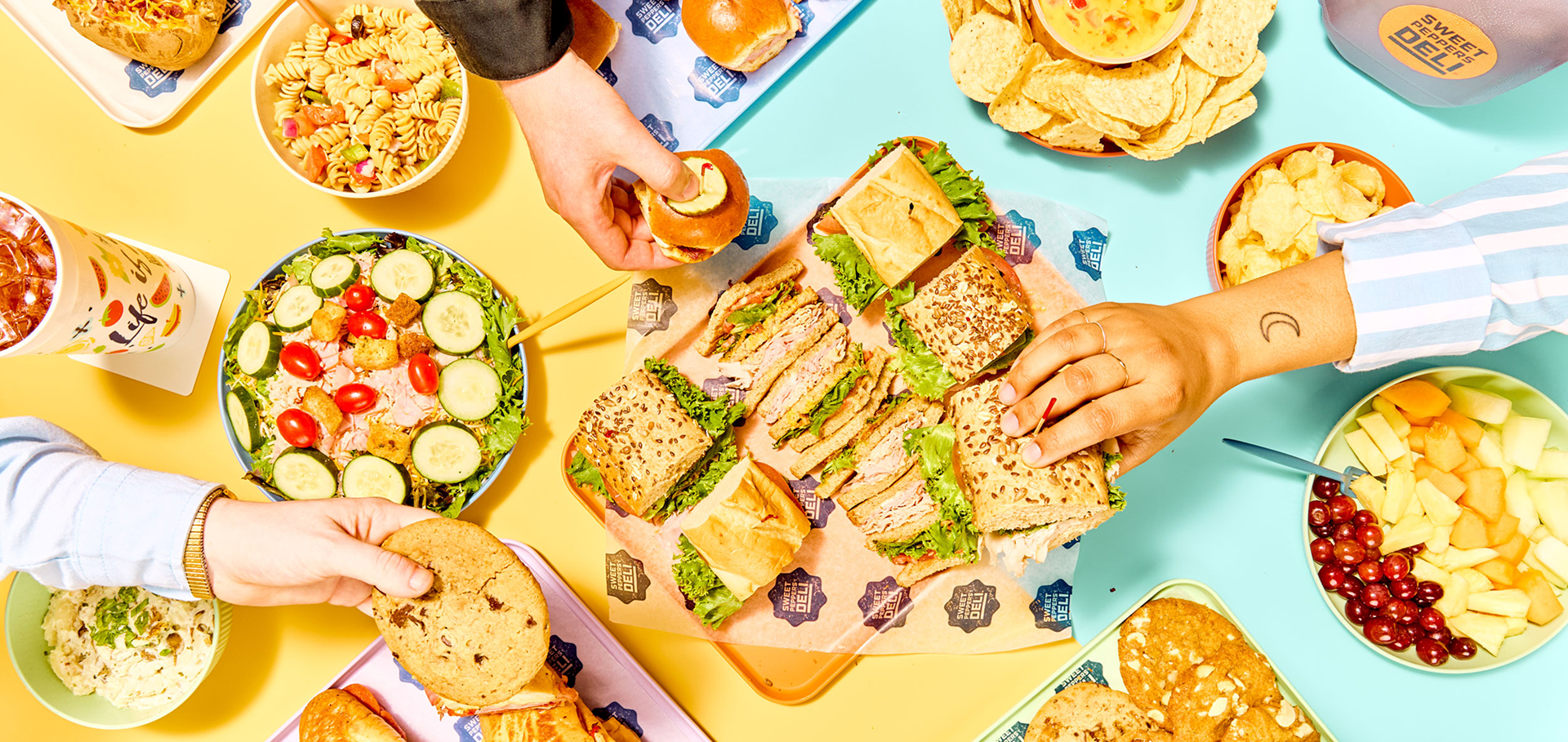table with hands holding sandwiches, cookies, chips, and sweet tea